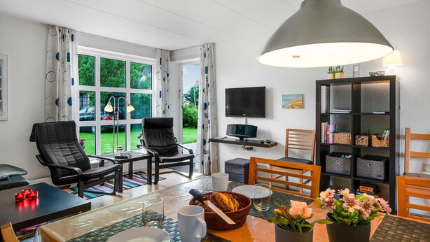 Un salon moderne avec un coin repas, comprenant une grande fenêtre, un téléviseur à écran plat, une bibliothèque et une table à manger agrémentée d’un panier de pain et de fleurs.