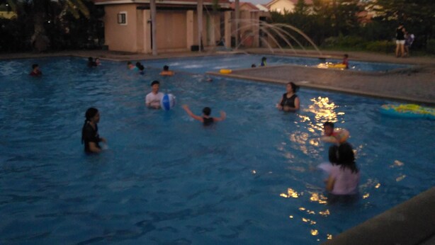 A swimming pool with people enjoying the water, surrounded by palm trees and a building with a red roof. - San Fernando Pampanga (Sanfernando)