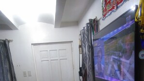 A room with a television mounted on the wall displaying a program, a standing fan, and a door with a patterned curtain. - San Fernando Pampanga (Sanfernando)