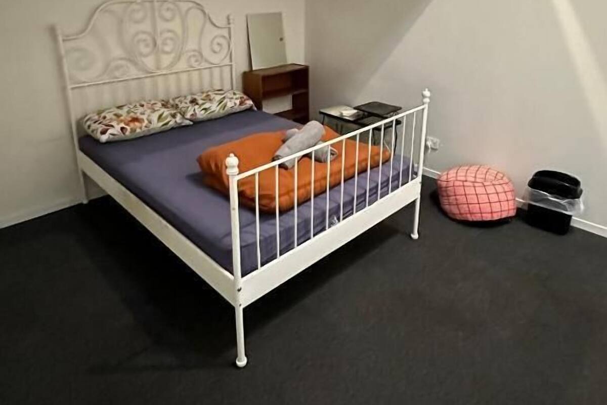 Quarto com estrutura de cama em metal, uma pequena mesa de cabeceira, um espelho e um pufe vermelho.