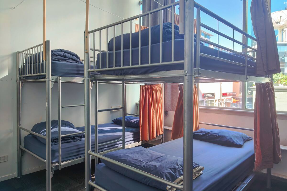 Quarto com beliche, grades de metal, roupa de cama azul e cortinas laranja.