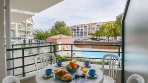 Een balkon met een ontbijttafel, compleet met croissants, fruit en sap, met uitzicht op een zwembad en woongebouwen.