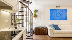 Una sala de estar moderna con un sofá blanco, un cuadro abstracto azul, una planta colgante y una maleta antigua.