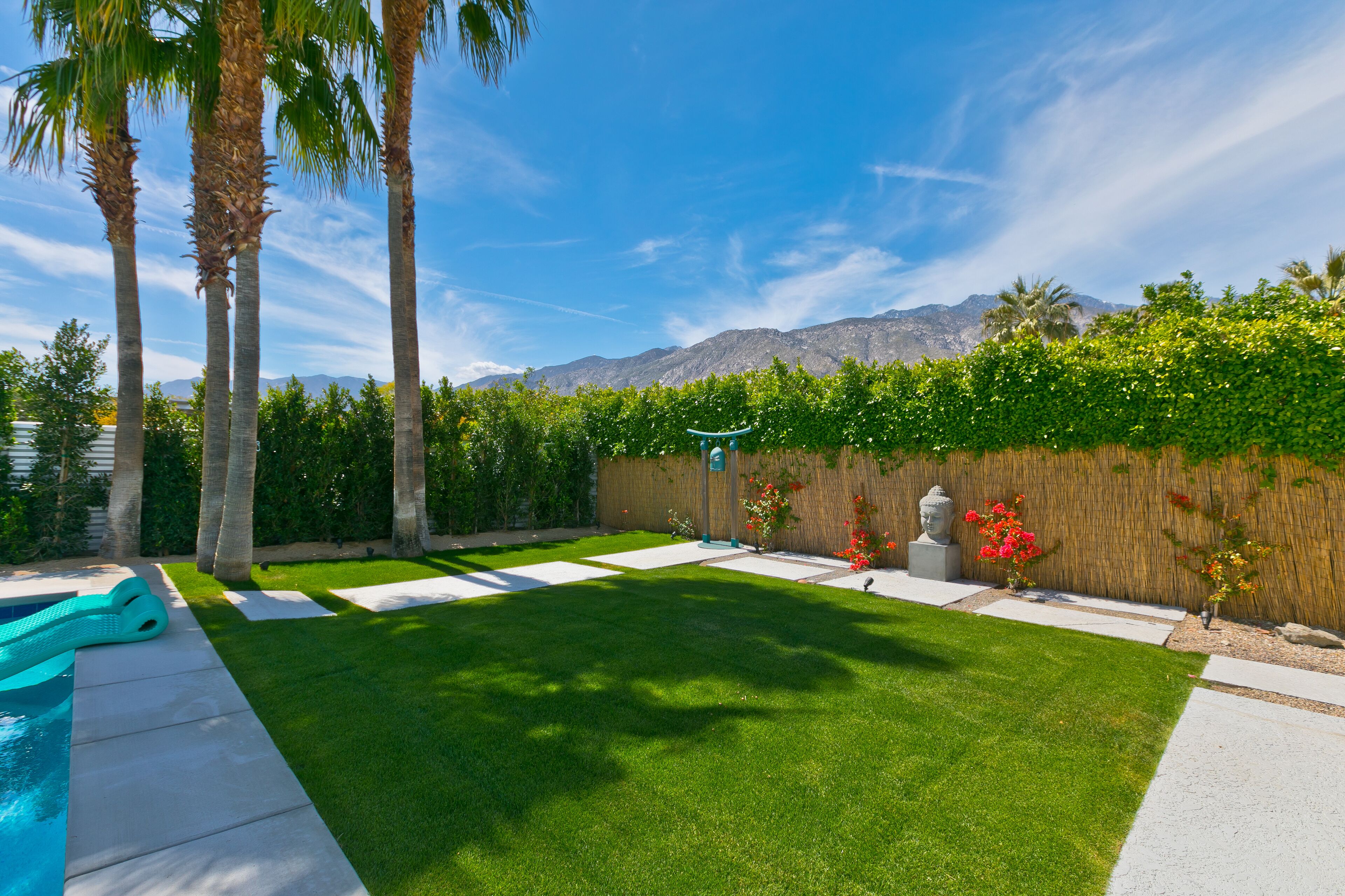 A backyard with a swimming pool, palm trees, a wooden fence, and a mountain view.