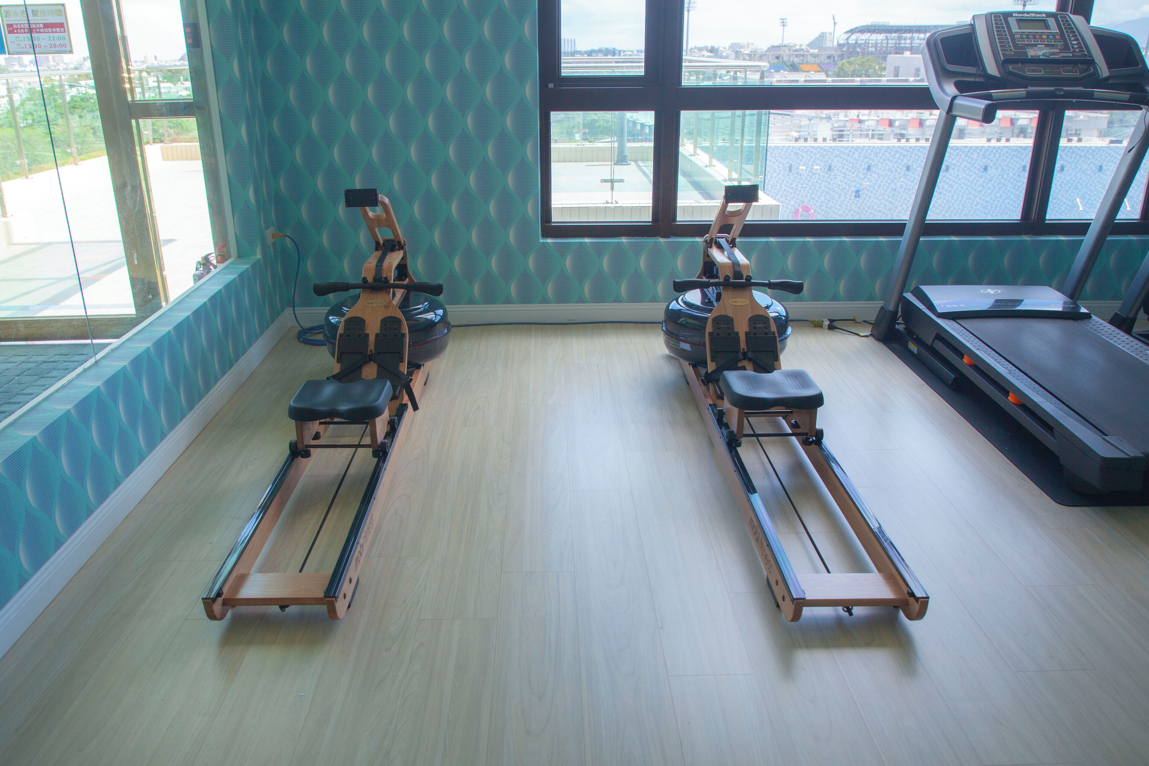 A gym with two rowing machines, a treadmill, and large windows with a view of a stadium.