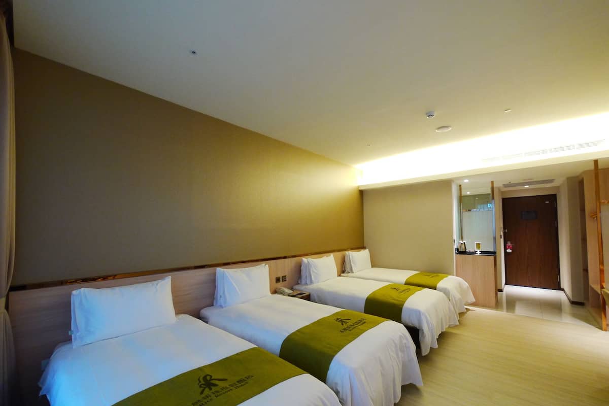 Habitació d'hotel amb tres llits, cadascun amb roba de llit blanca i estovat verd. Hi ha un capçal de llit de fusta, una taula de nit amb una làmpada i una porta que dona a una altra habitació.