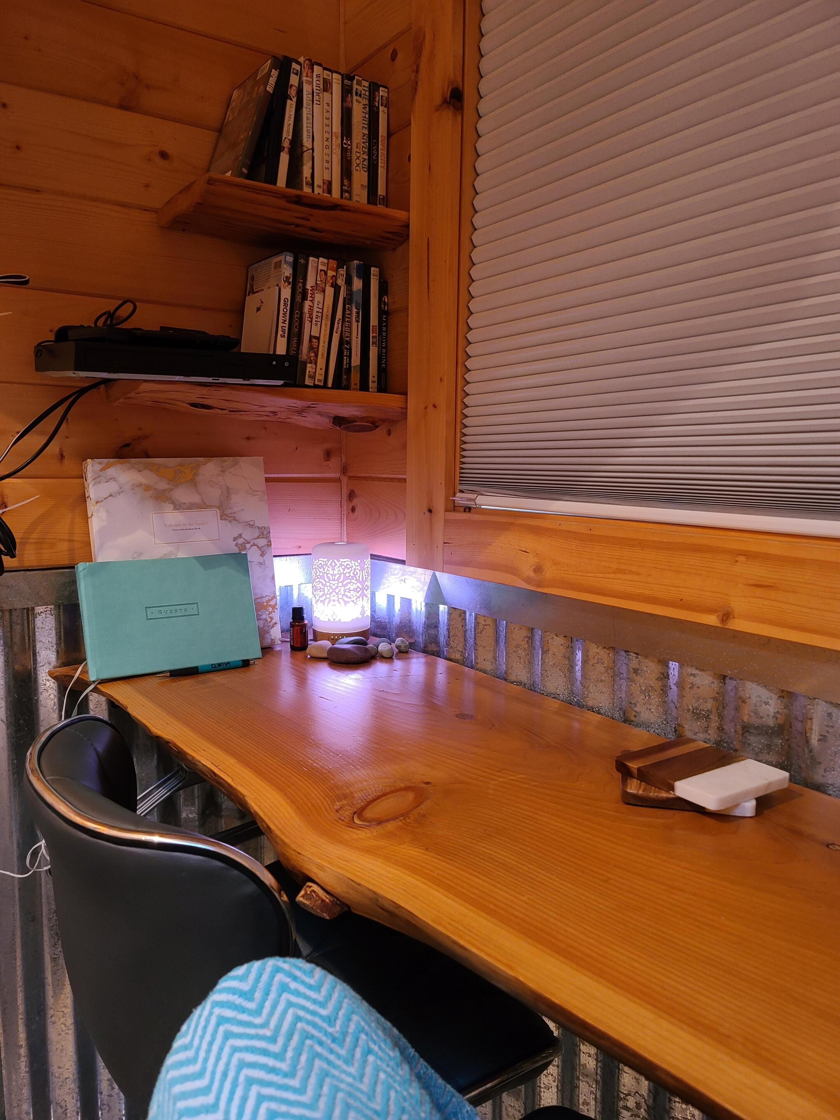 A wooden cabin interior with a desk, bookshelves, a window with blinds, and a chair.