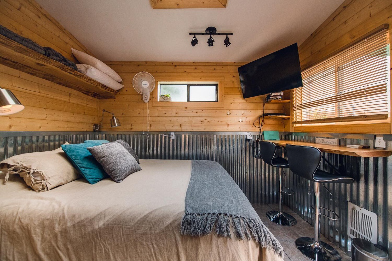 A cozy room with a bed, a desk, a chair, a TV, and a window with blinds.