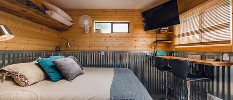 A cozy room with a bed, a desk, a chair, a TV, and a window with blinds.