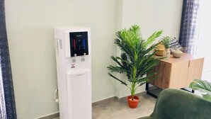 Un distributeur d’eau, une plante en pot et un meuble en bois dans une pièce aux murs de couleur claire et au sol carrelé.