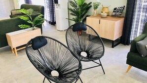 Un salon moderne avec un canapé vert, deux fauteuils à structure métallique noire, une table d’appoint en bois et un meuble contenant des objets décoratifs.