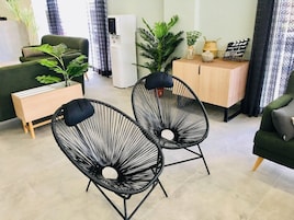 A modern living room with a green sofa, two black wireframe chairs, a wooden side table, and a cabinet with decorative items.