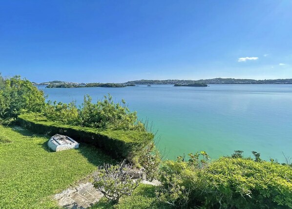 A coastal landscape with a grassy area, a stone structure, and calm water with distant islands.