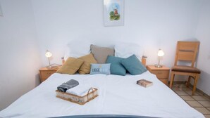 Ein ordentlich bezogenes Bett mit weißer Decke, mehreren Kissen, einem kleinen Nachttisch mit Lampe und einem Stuhl.