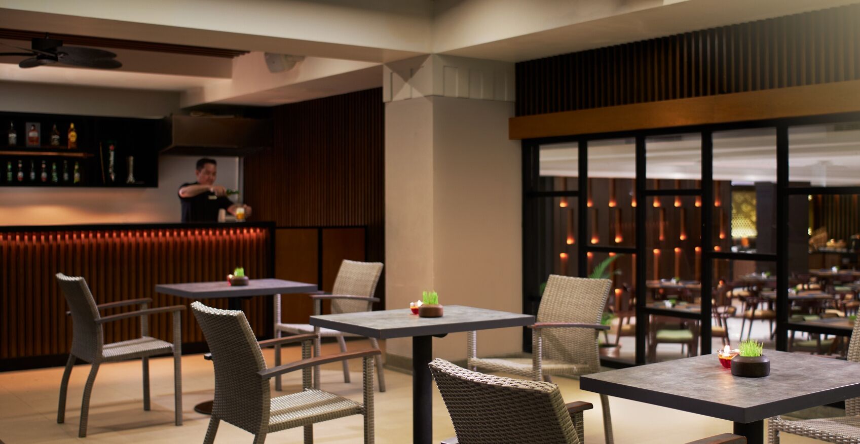 Un interno ristorante moderno con sedie e tavoli in vimini, una zona bar e ampie finestre che lasciano entrare la luce naturale.