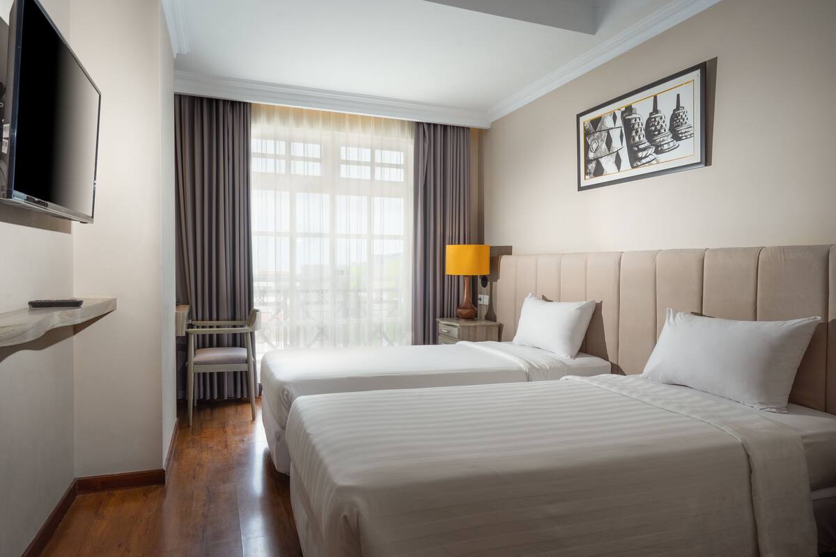 Une chambre d’hôtel avec deux lits, une télévision, une fenêtre avec des rideaux, un tableau encadré et une table de chevet avec une lampe.