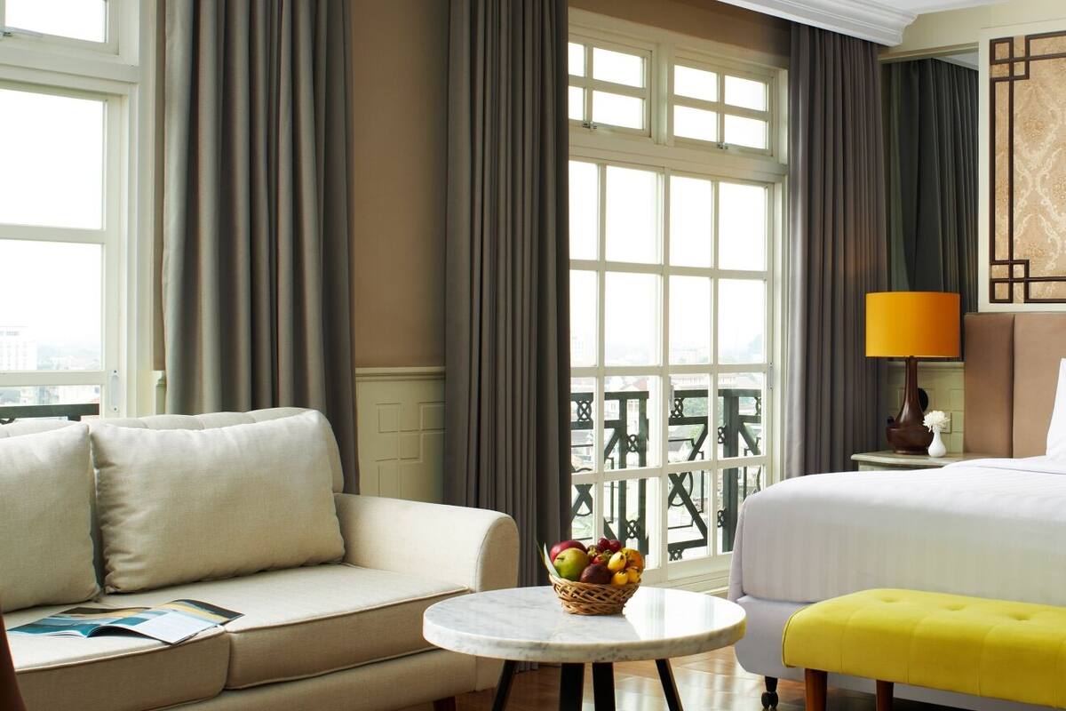 Une chambre d’hôtel avec un lit, un canapé, une petite table ronde sur laquelle se trouve un panier de fruits, et une fenêtre avec des rideaux.