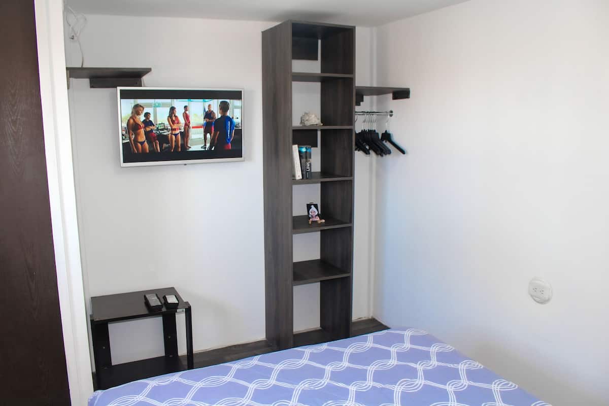 A bedroom with a bed, a TV mounted on the wall, a small table, and a shelving unit with items on it.