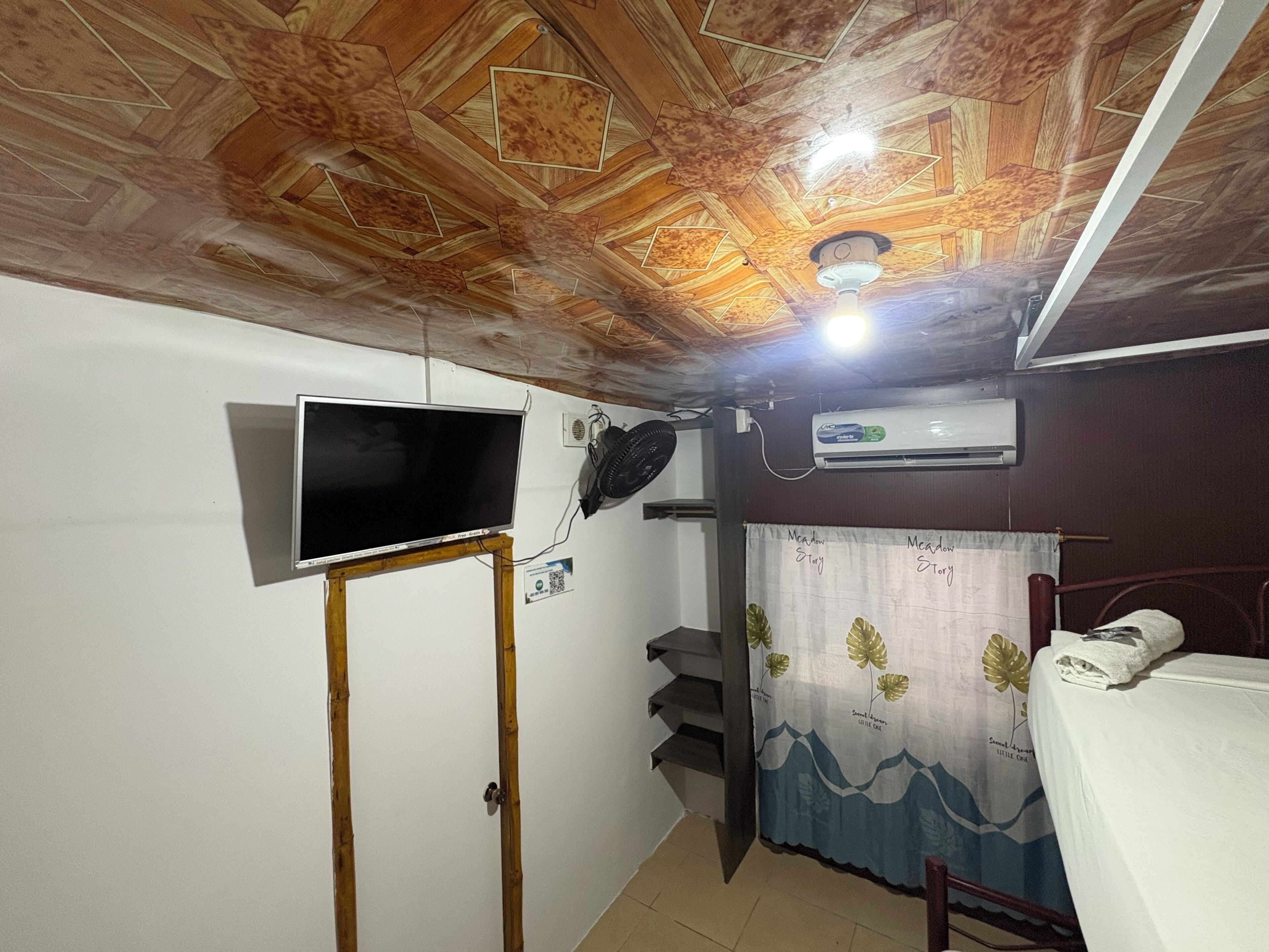 A room with a wooden ceiling, a mounted TV, an air conditioner, a bed, and a curtain with a mountain design.