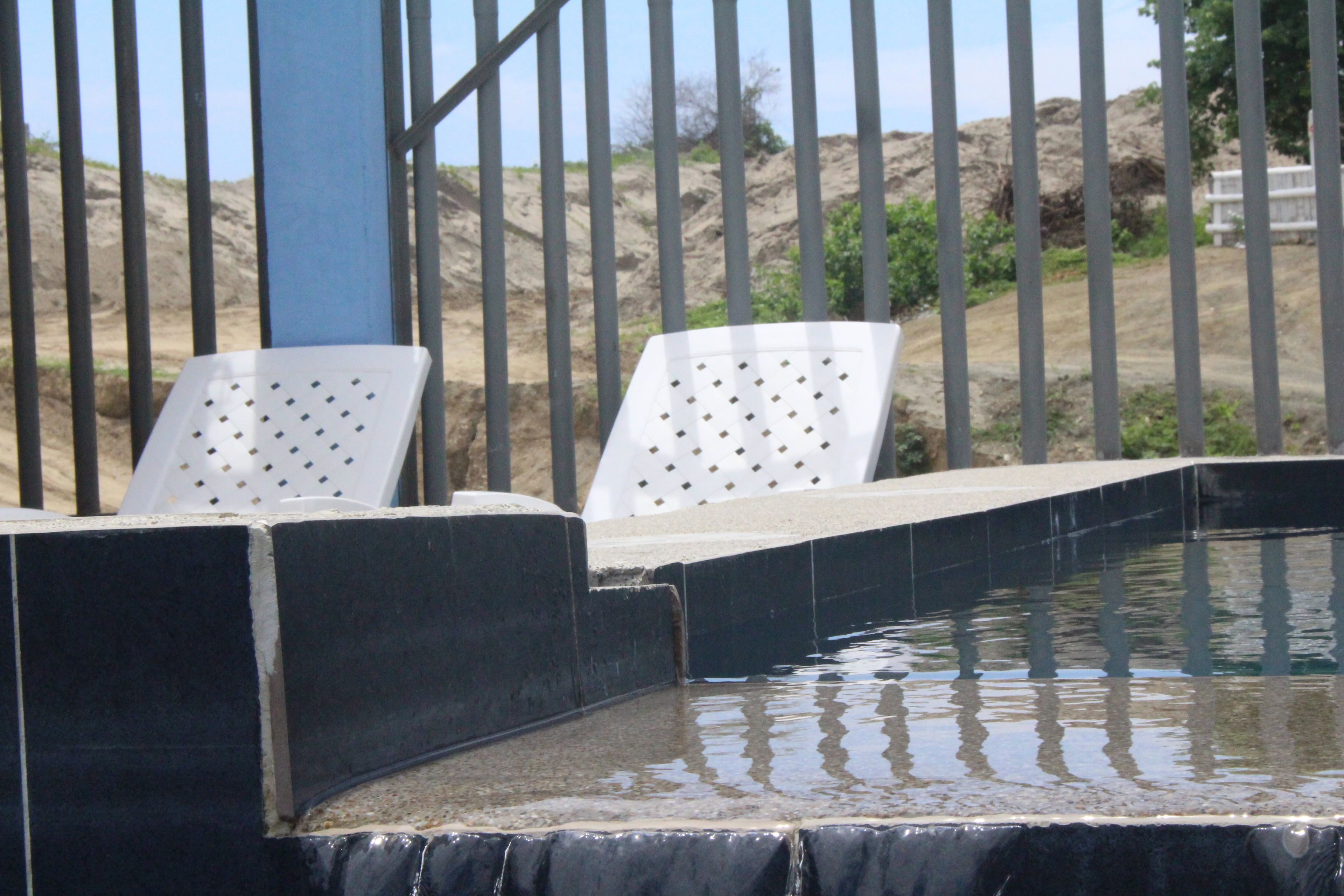 Zona de piscina con escalones que bajan al agua, rodeada por una valla metálica y sillas de descanso blancas.