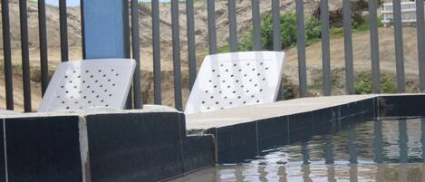 Zona de piscina con escalones que bajan al agua, rodeada por una valla metálica y sillas de descanso blancas.