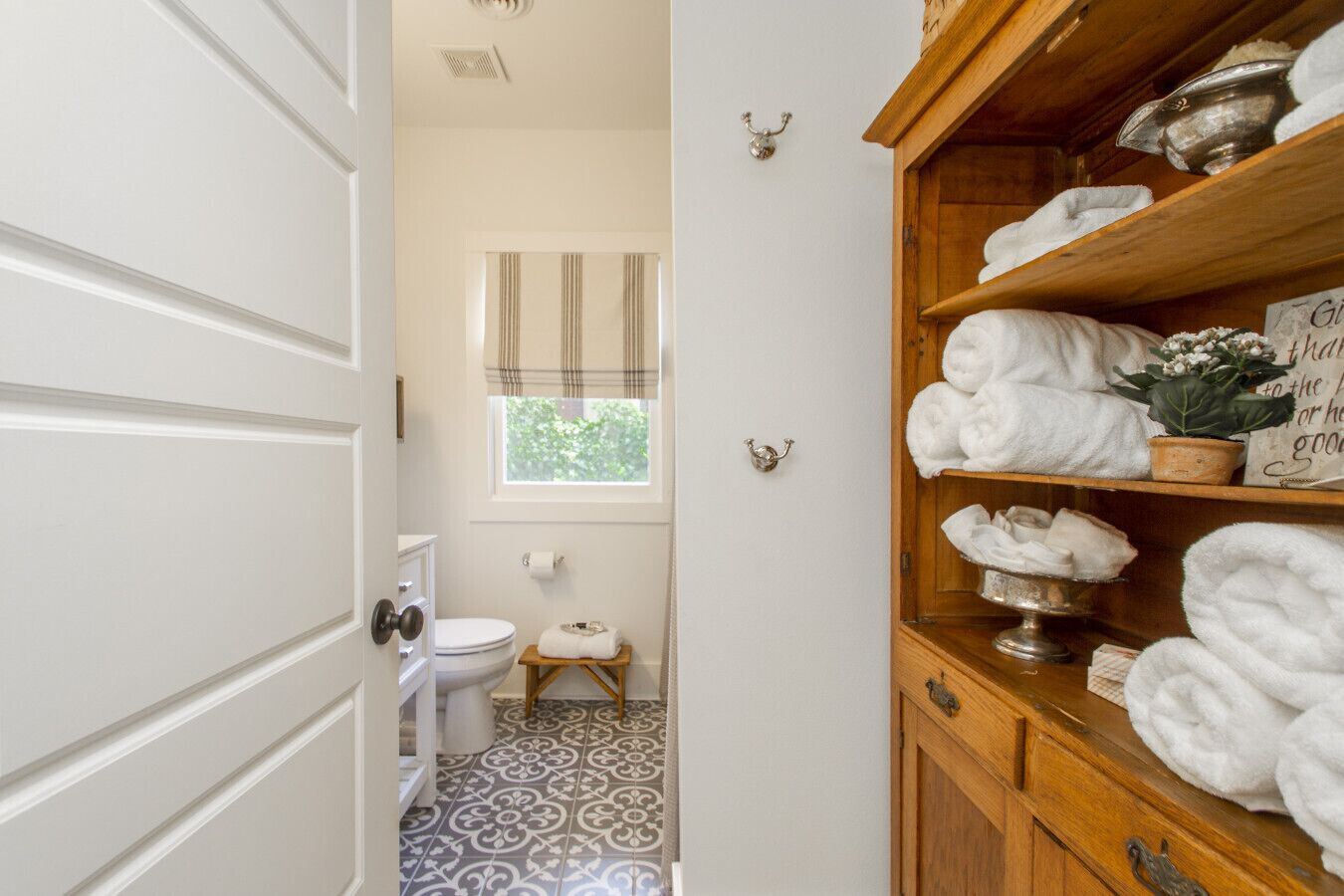 Une salle de bain avec des toilettes, un petit tabouret en bois, une fenêtre avec des rideaux et une étagère avec des serviettes.