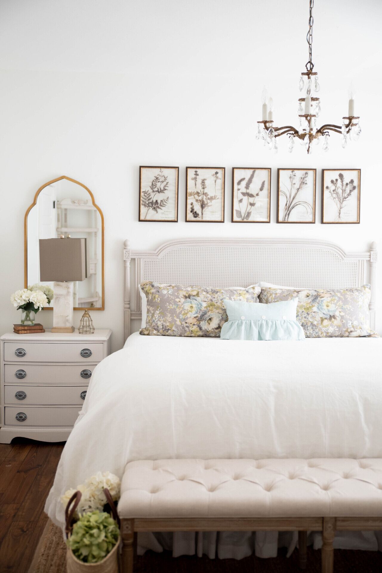 A bedroom with a white bed, floral pillows, a bench, a nightstand with a lamp, a mirror, and framed botanical prints on the wall.