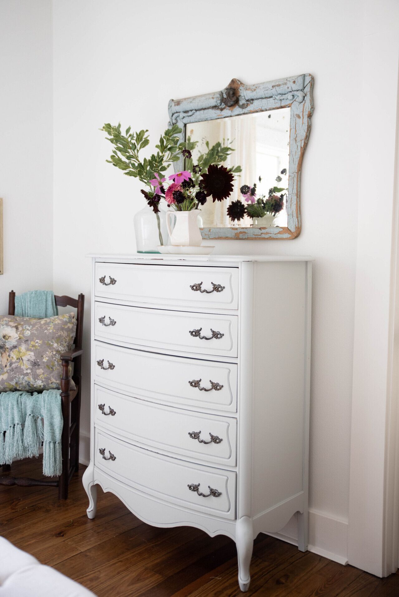 A white dresser with ornate handles, a mirror with peeling paint, and a vase with flowers on top.