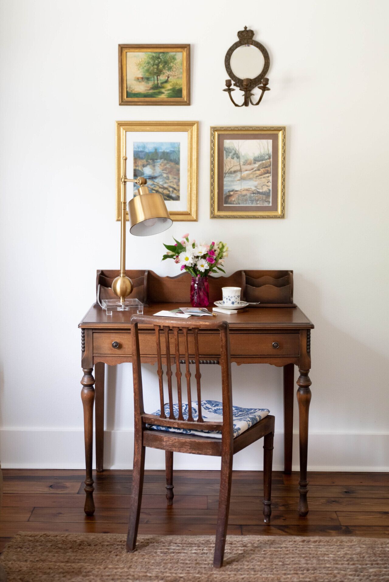 Een houten bureau met een lamp, een vaas met bloemen en ingelijste foto’s aan de muur.