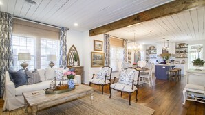 A spacious living room with a wooden floor, a white sofa, and a dining area with a chandelier. - Charming Historic District Cottage Near Downtown (Conway)