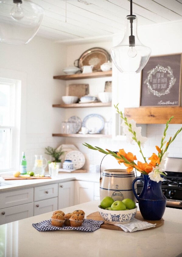 Una encimera de cocina con un florero azul, un bol de manzanas verdes y magdalenas sobre un mantel a cuadros.