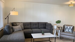 Ein modernes Wohnzimmer mit einem Ecksofa, einem weißen Couchtisch, einem kleinen Beistelltisch mit Pflanze und einem Sessel mit gemustertem Kissen.