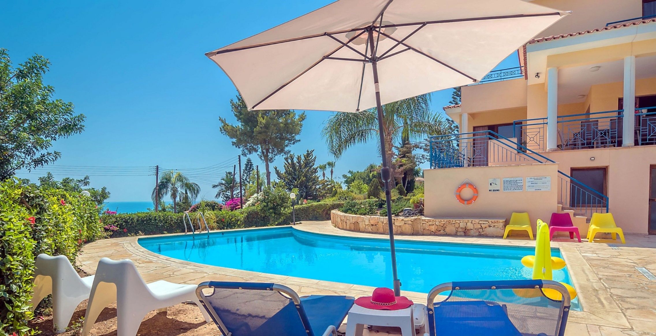 Una piscina con sillas de descanso y sombrillas, rodeada de vegetación y un edificio con balcones.