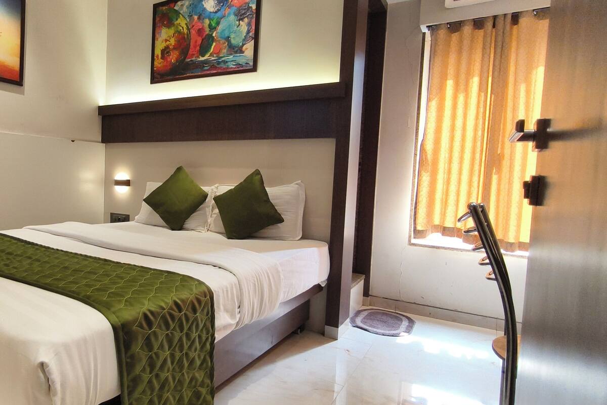 Una habitación de hotel con cama, mesita de noche, lámpara de pared y ventana con cortinas.