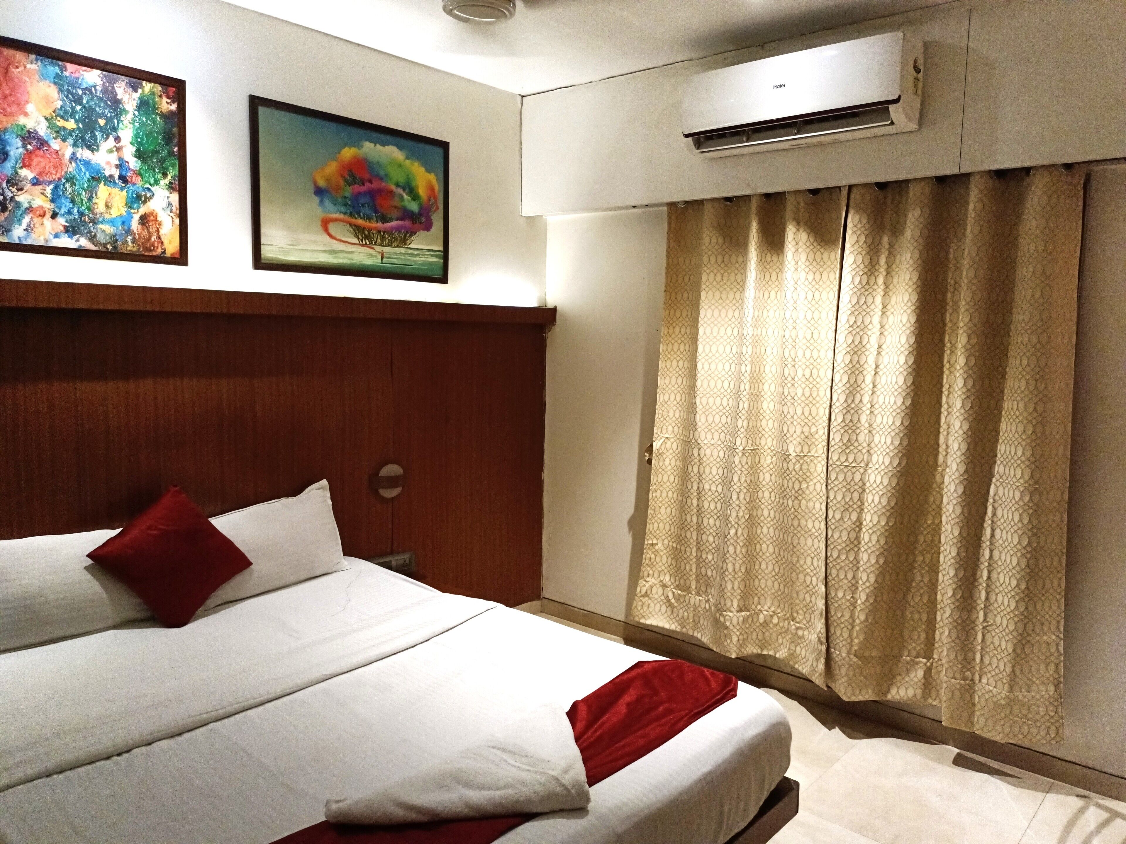A hotel room with a bed, a wooden headboard, a red pillow, a white bedspread, a red blanket, a window with a curtain, and an air conditioning unit.