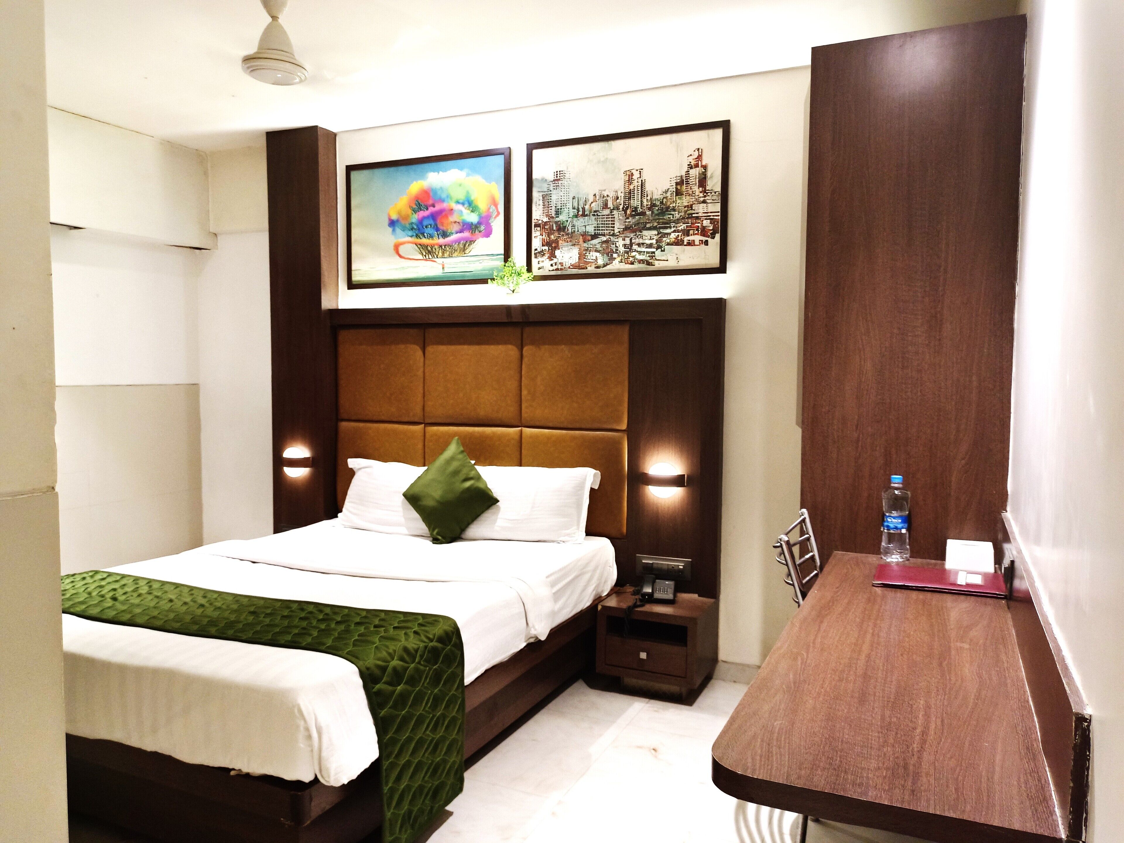 A hotel room with a bed, a nightstand, a desk, and two framed pictures on the wall.