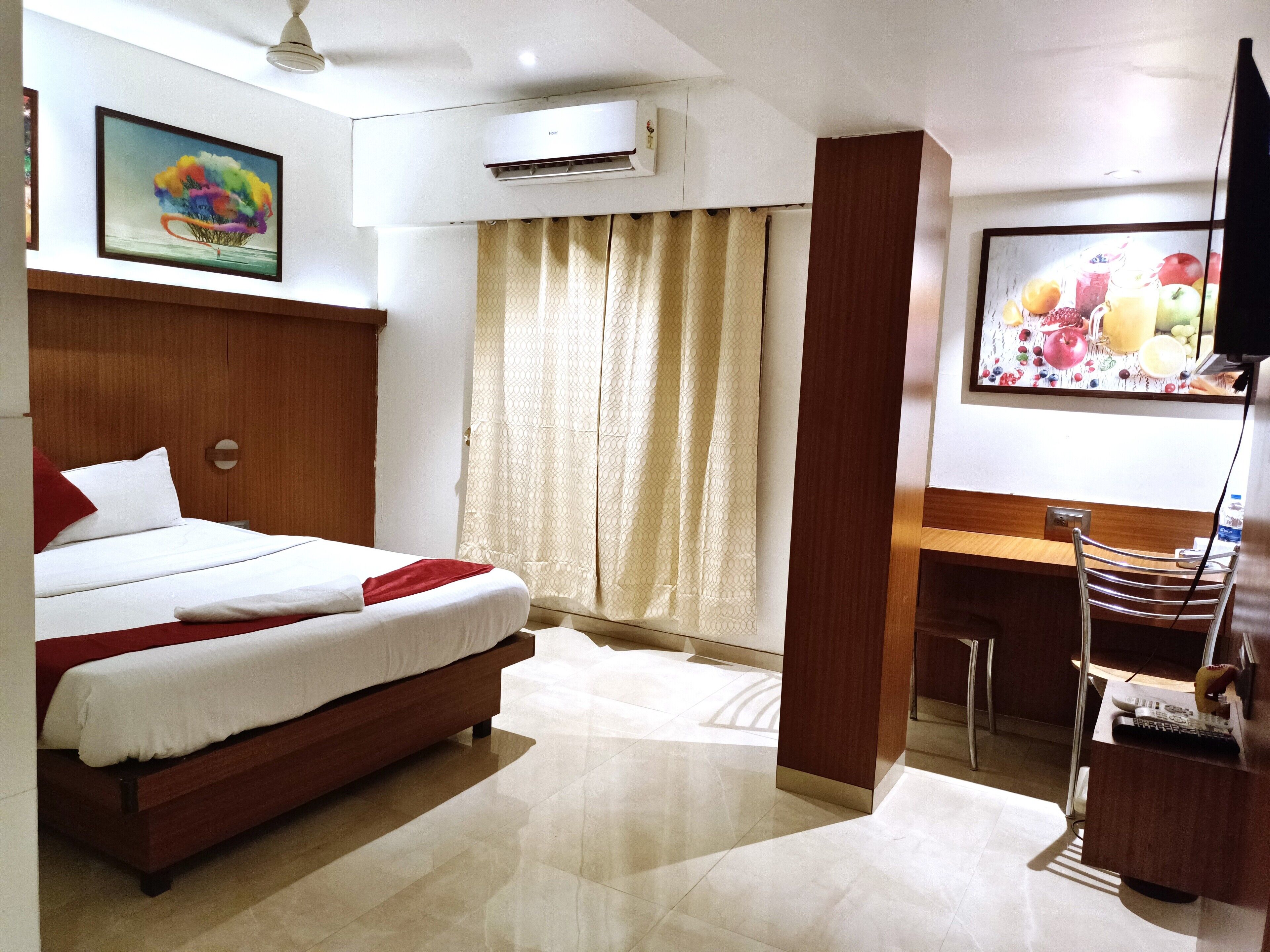 A hotel room with a bed, a desk, a chair, an air conditioner, a TV, and a painting on the wall.