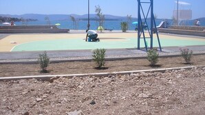 Un terrain de basket avec un panier bleu, une zone sablonneuse et une étendue d’eau en arrière-plan.