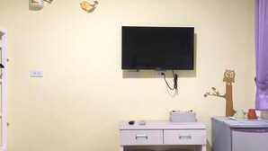 Una habitación con televisión montada en la pared, un escritorio con dos sillas, un cubo de basura y un refrigerador pequeño.