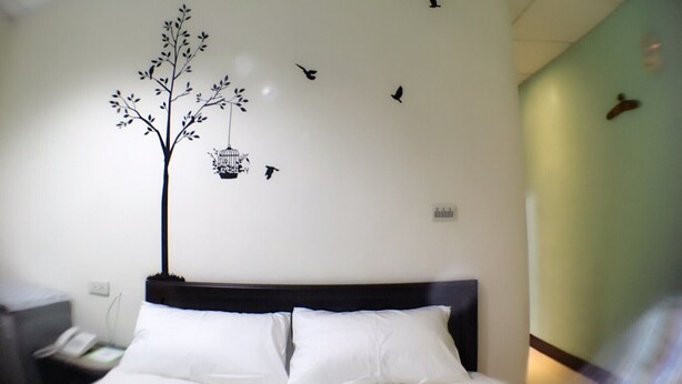 Habitación de hotel con cama, mesita de noche, pared decorada con un adhesivo de árbol y un pájaro, e interruptor de luz.