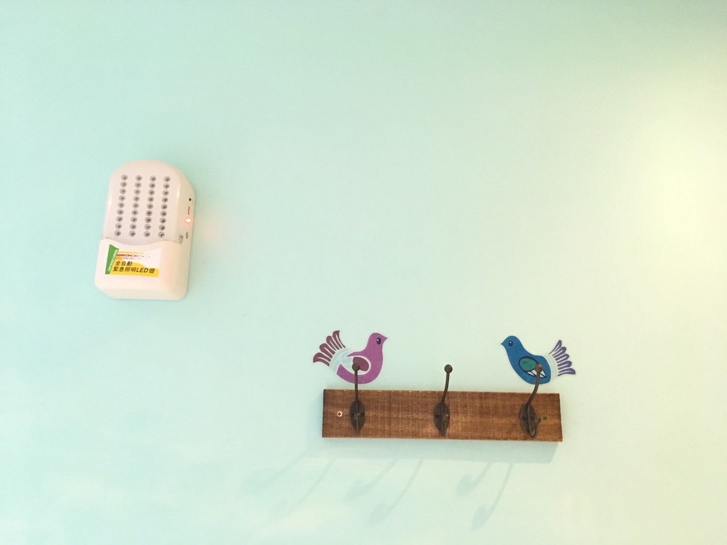 Un estante de madera fijado a la pared con tres ganchos con forma de pájaro, una luz LED rosa y un pequeño objeto blanco con texto.