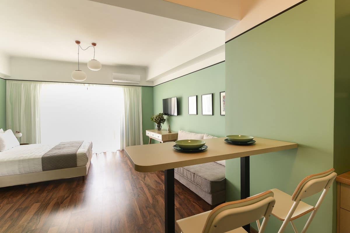 A modern hotel room with a large bed, a dining table with chairs, a small sofa, and a television on the wall.