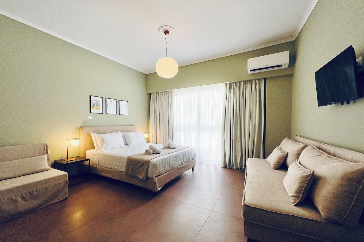 A hotel room with a bed, two sofas, a nightstand with a lamp, a ceiling light, a window with curtains, and an air conditioning unit.