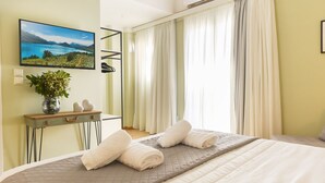 A hotel room with a bed, a TV mounted on the wall, a desk with a vase and decorative items, and a window with curtains. - Athens Stories (Athens)