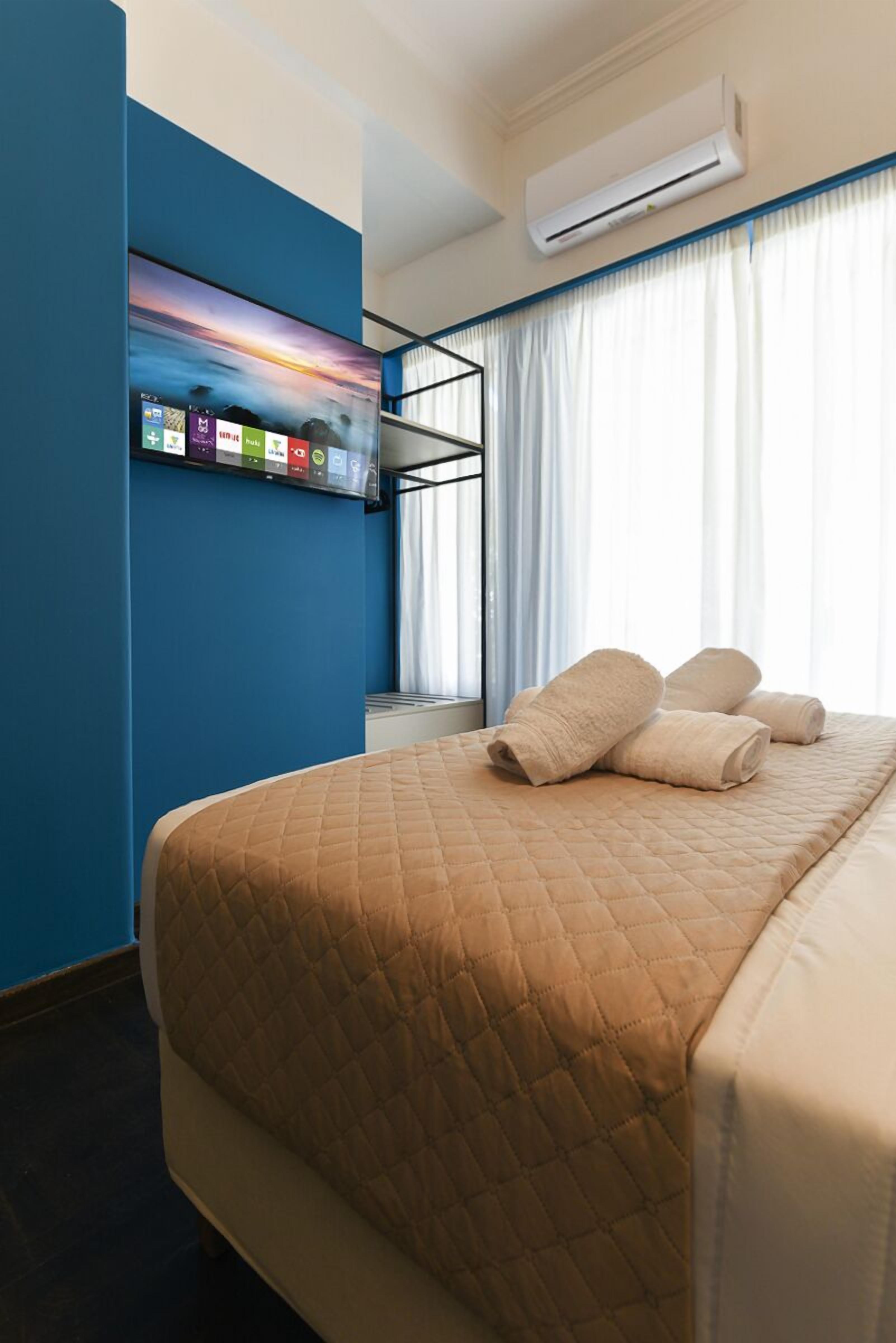 A hotel room with a bed, a wall-mounted TV, and an air conditioning unit.