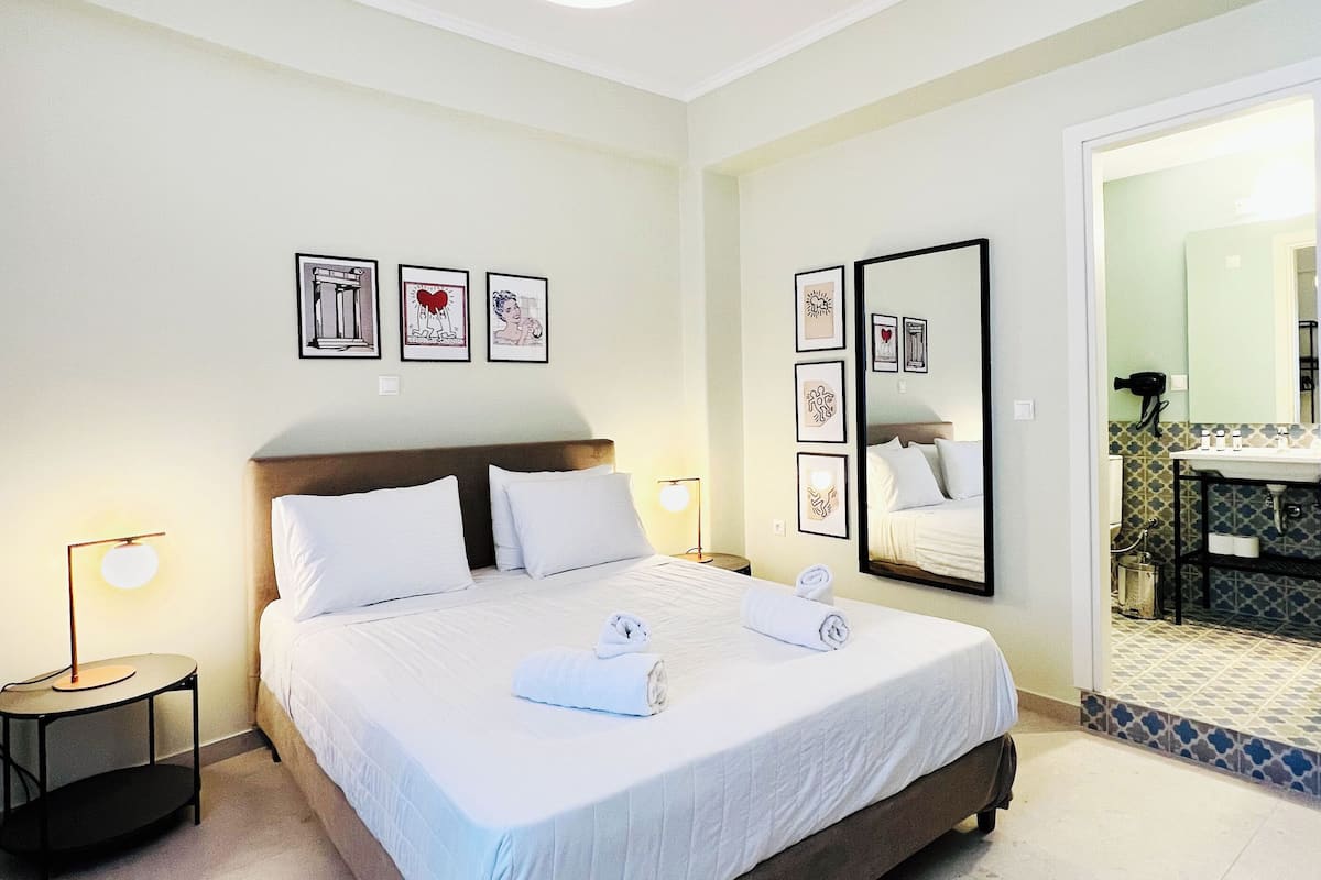 A neatly made bed with white linens and two rolled towels, a bedside table with a lamp, and a wall with framed artwork.