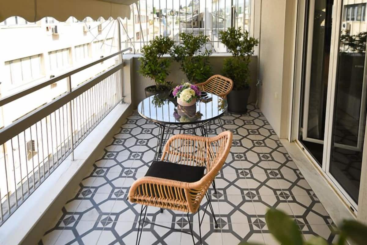 A balcony with a patterned floor, wicker chairs, a glass table, and potted plants.