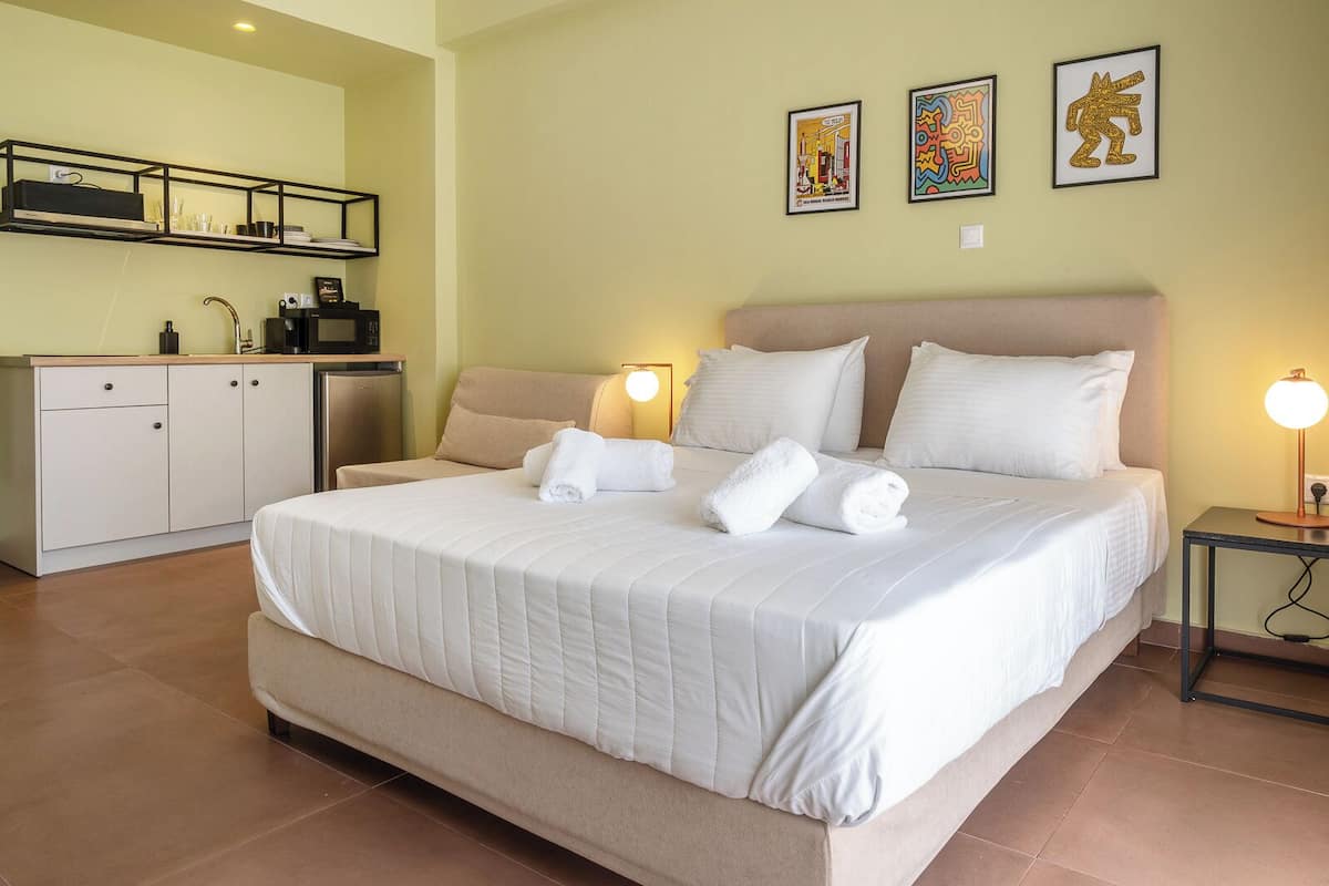 A hotel room with a bed, bedside lamps, a small table, a desk with a chair, and artwork on the wall.