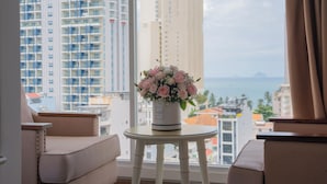A hotel room with a view of tall buildings and the sea. There is a white table with a vase of flowers and a chair. - Nha VIET Nha Trang Hotel (Nha Trang)