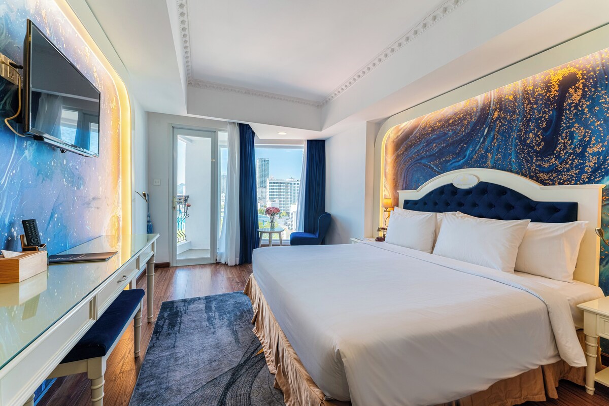 Ein Hotelzimmer mit einem großen Bett, einem an der Wand befestigten Fernseher, einem Schreibtisch mit einer Bank und Blick auf die Stadt durch das Fenster.